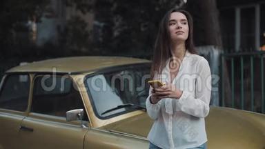 年轻迷人的布鲁内特白种人女孩站在老时尚的<strong>金色汽车</strong>边使用她的智能手机和